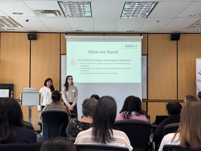 Image of members Ava and Saanya giving a presentation in front of an audience, the slide showing "What we found" under "social relationship and networks", and "family or home-care settings supported social relationships and engagement. Staffing limitations and peer conflict created barriers to positive interactions. Lack of support increased risk of social isolation"