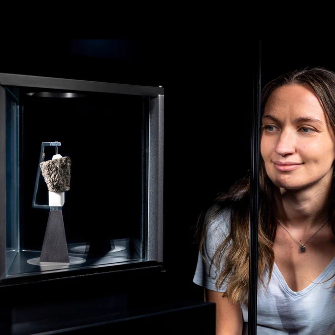 A Science Museum visitor looks closely at a grey rock from the moon in a glass case in the Space gallery. The background is dark and the moon rock well-lit in its case. 