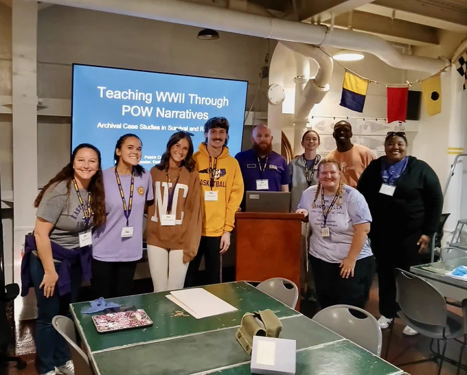 A group of people standing in front of a a presentation slide titled "Teaching WWII Through POW Narratives" with the subtitle "Archival Case Studies in Survival and Rescue." The room has nautical flags hanging from the ceiling. 
