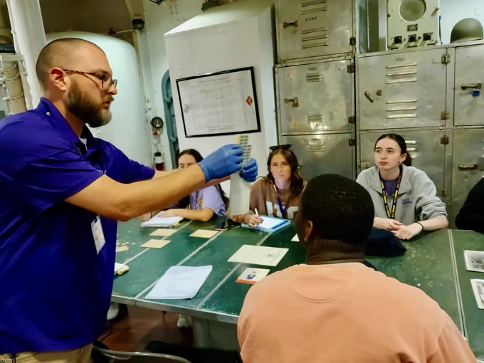 A person wearing gloves holds items in the air demonstrating to a group of people seated around a table. 