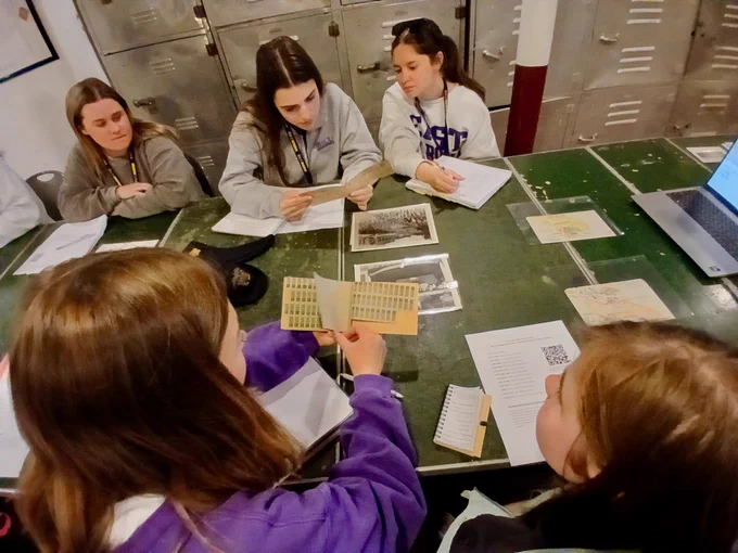 A group of people sitting around a table covered with papers, a laptop, and photographs, engaged in discussion. They're in a room with metal lockers. One person holds a sheet with a grid pattern.