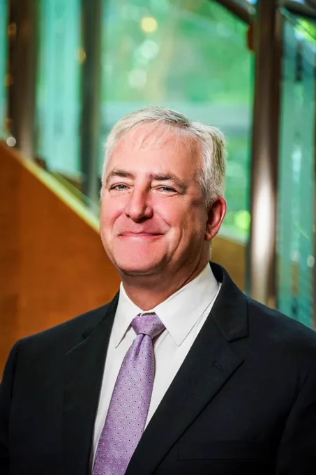 Headshot of Rob Puentes, the new CEO of the Lincoln Institute of Land Policy, smiling and wearing a dark suit, white shirt, and light purple tie, standing indoors in front of large windows with greenery visible outside.