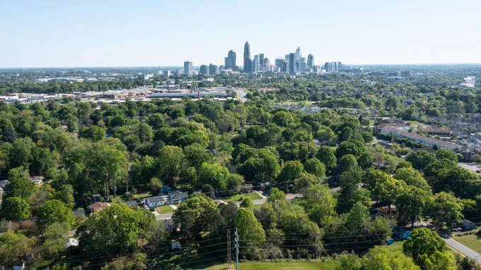 Aerial view of a forested suburban area with scattered houses in the foreground. In the distance, the skyline of Charlotte, North Carolina is visible, featuring tall buildings under a clear blue sky.