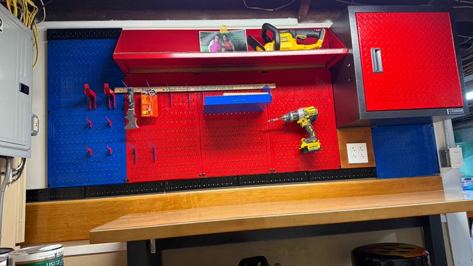 Organized workshop featuring red and blue Wall Control metal pegboards mounted above a wooden workbench, holding a drill, hand tools, hooks, and storage accessories, with a matching red cabinet and neatly arranged workspace.