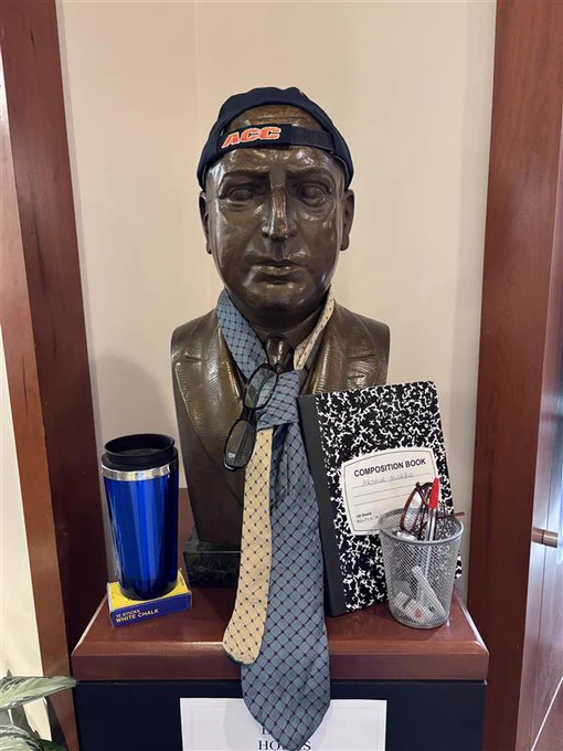 A bust of Arthur J. Morris wearing a backwards baseball cap and a loose tie. He is surrounded by a coffee cup, a notebook, and a cup of pens and glasses.