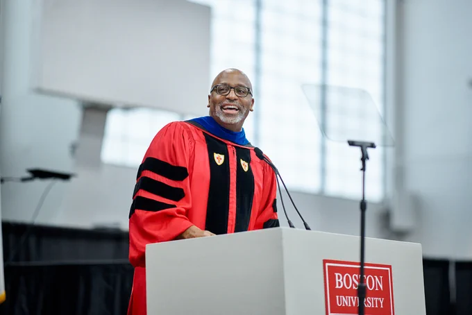 A person wearing a red doctoral gown with black velvet chevrons and a blue hood stands behind a white podium labeled \"Boston University,\" speaking into a microphone; a clear speech shield and large windows blur in the bright background.