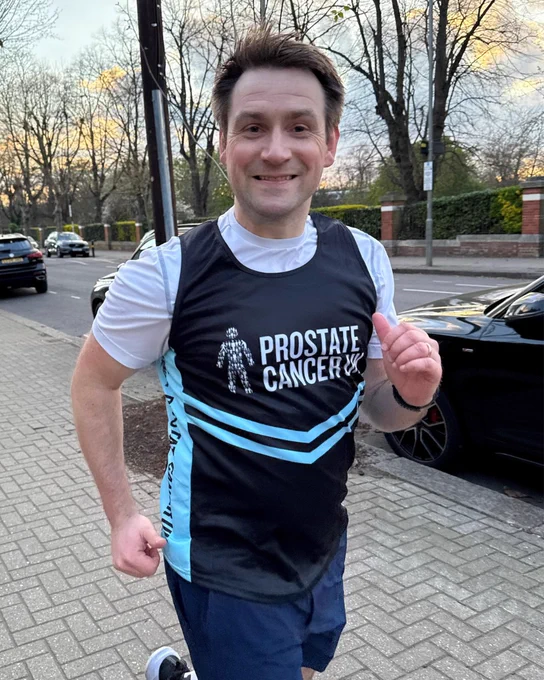 Alex jogging on a pavement wearing a Prostate Cancer UK running vest. Trees and parked cars line the street in the background.