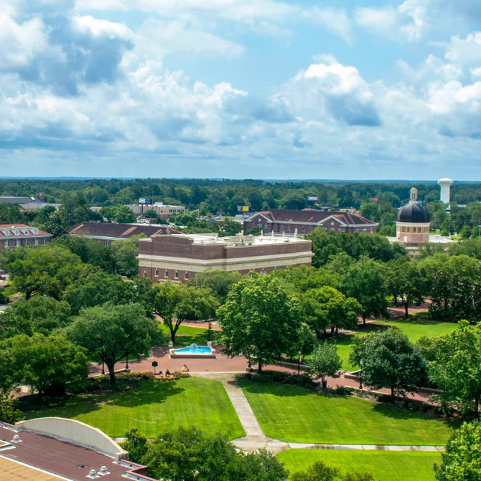 Aerial view of the USM Hattiesburg campus. Sidewalks are seen among green lawns, trees, and brick buildings. The Aubrey K. Lucas Administration building can be seen, along with the fountain at Shoemaker Square. A blue sky is overhead.
