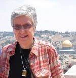 Photograph of Mary Gray wearing sunglasses and a red plaid collared shirt on top of a black shirt with a necklace, and standing outdoors with arid hills and buildings in the background.