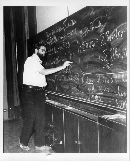 Photograph of Alfred (Alfie) Gray wearing glasses, a white shirt, and black pants standing at a blackboard writing mathematics with chalk.