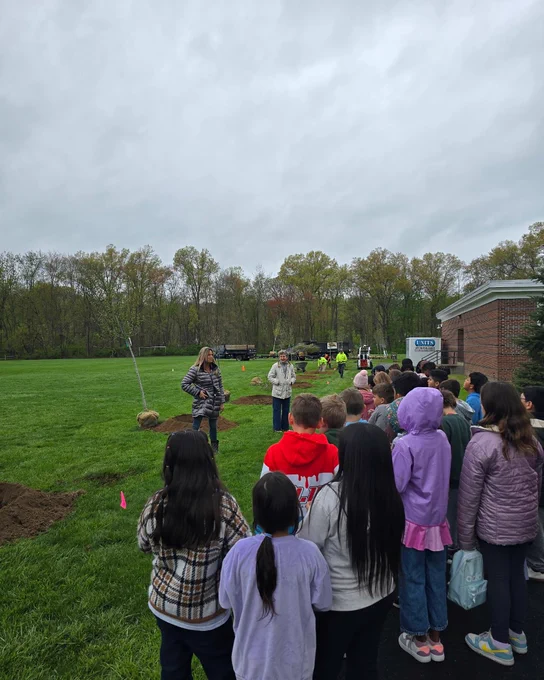 A group of students standing on a grassy field, with a woman addressing them. Trees and a building are visible in the background.