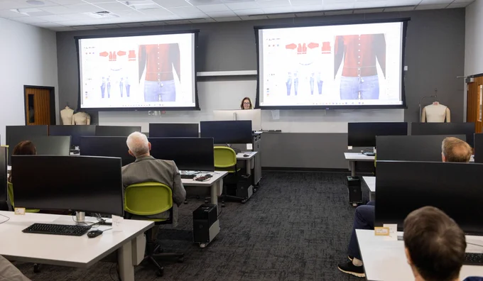 A classroom with several people seated at desks facing forward. Two large screens display fashion design software featuring clothing patterns. A person is standing at the front, near the screens, giving a presentation. The room has modern furnishings with multiple computer monitors and chairs.