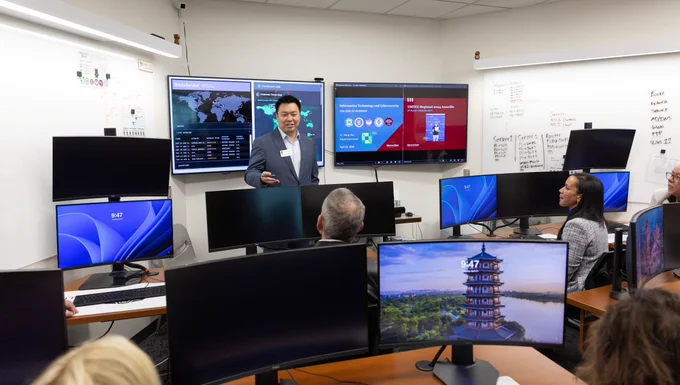 A person is presenting in a control room with multiple computer screens. The room includes monitors displaying world maps, graphs, and a pagoda image. The audience is seated at desks.