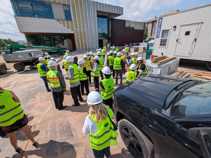 A group of people wearing safety vests and helmets are gathered in a construction site where a large building is partially constructed. A trailer with "Milis" written on it is visible, and several construction vehicles are present.