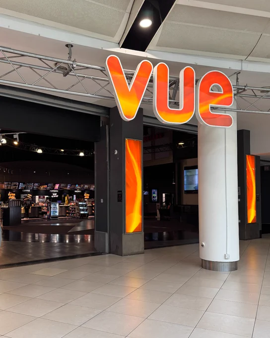 Entrance of a Vue cinema, featuring an illuminated "Vue" sign. Inside, there are visible concession stands and digital screens.