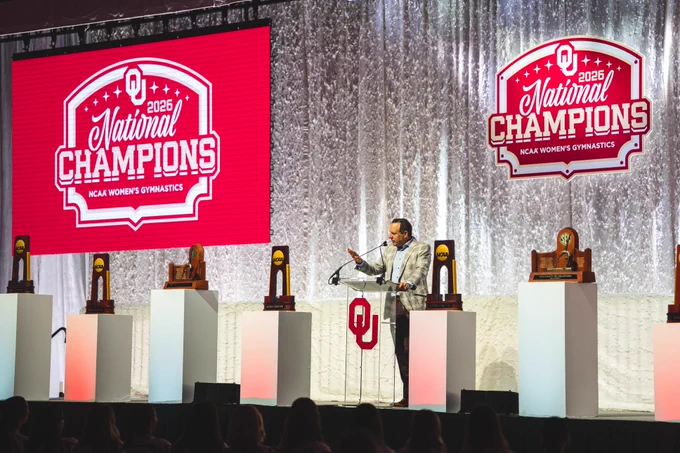 President Joseph Harroz Jr. stands at a podium on a stage with a large "OU 2026 National Champions" sign displayed. Trophies are arranged on white pedestals around the stage, adorned with the OU logo.