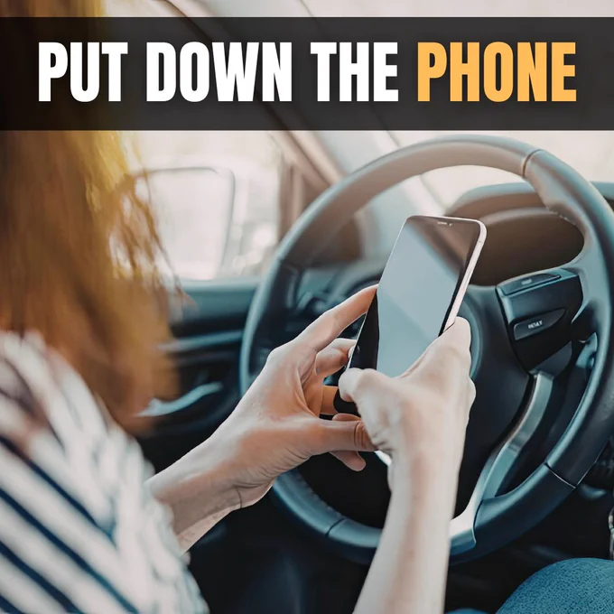 A person sitting in a car is holding a smartphone with both hands, placed in front of a steering wheel. A text overlay reads, "PUT DOWN THE PHONE."
