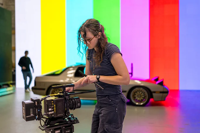 A student with long wavy hair stands in a sound stage space while adjusting camera equipment. There is a prop car and a large screen behind them. 