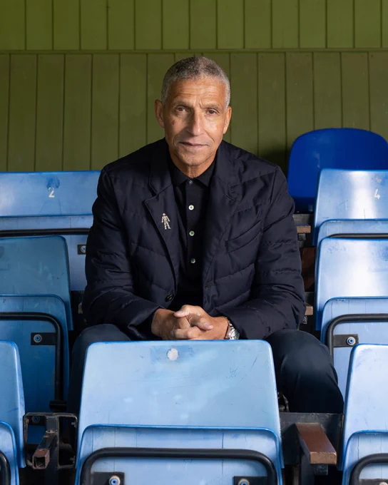 Chris Hughton is sitting in a row of empty blue stadium seats wearing a black jacket.
