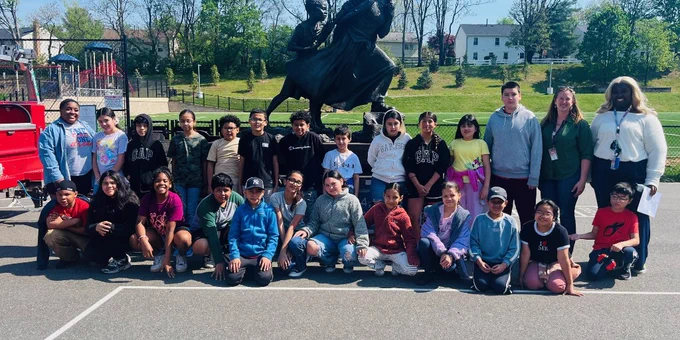 Group of diverse children and adults posing outdoors in front of a large statue on a sunny day.