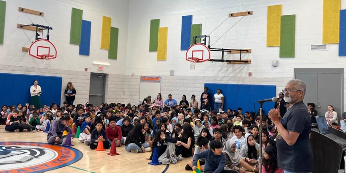 A large group of children seated on a gymnasium floor listening Anthony Cohen with a microphone near a basketball hoop.