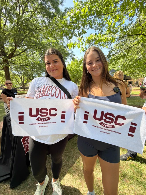 Two individuals hold up towels with "USC Alumni" written on them. They are outdoors, surrounded by trees and other people in the background. One person wears a shirt with "Gamecock" printed on it.