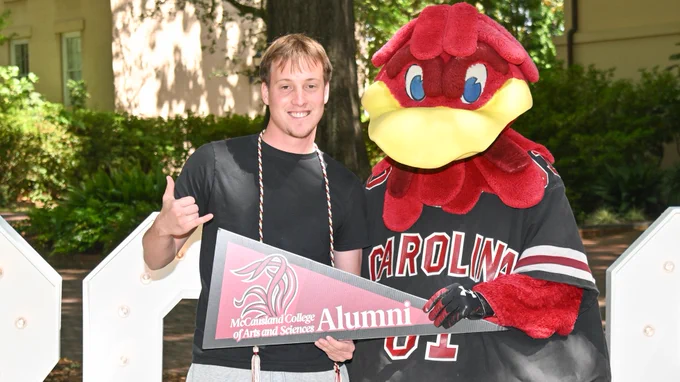 A person stands next to the University of South Carolina mascot, Cocky, holding a McCausland College of Arts and Sciences alumni pennant. They are outdoors with large white light-up numbers in the background.