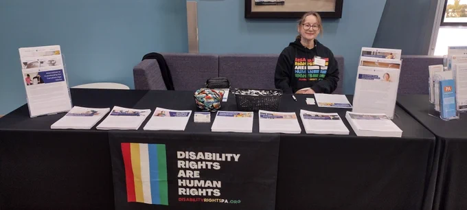 Photo of DRP Director of Intake and Grants Management, Shari Mamas, behind a standard DRP outreach table covered with resources, pens and pins. 