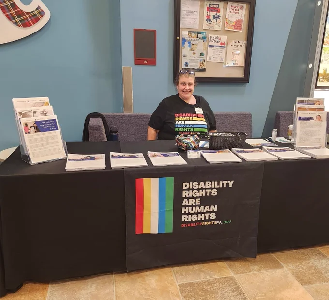 Photo of DRP's Administrative Coordinator, Stacey Mamas, behind a standard DRP outreach table covered with resources, pens and pins. 