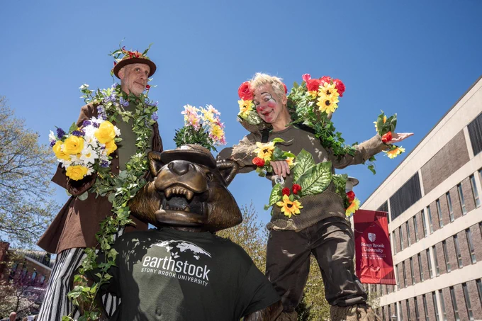 Two stilt performers dressed in nature-themed costumes with flowers and greenery stand beside the Wolfie Statue, wearing a "Stony Brook University Earthstock" shirt. The background includes a red "Stony Brook University" banner and the Library building.