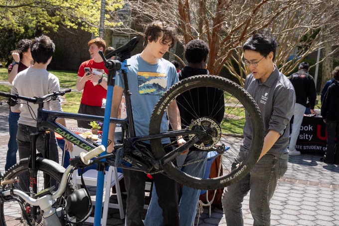 A group of people is gathered outside on the Academic Mall, focusing on a mountain bike mounted on a repair stand. A student is examining the bike, while others engage in conversation nearby. Trees and greenery surround the area.