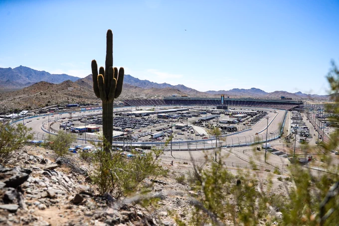 Panoramic view taken from the top of the hillside at Phoenix Raceway. You can see plants and a cactus in the foreground and cars racing around the track in the background.