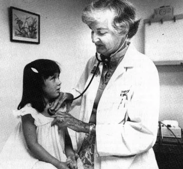 A medical professional uses a stethoscope to listen to a child's chest in an examination room. A framed botanical picture is visible on the wall.