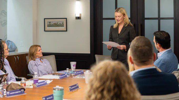 A person stands holding a paper, addressing a group seated around a conference table. The room has modern decor, with a framed picture on the wall. Various drinks and nameplates are on the table.