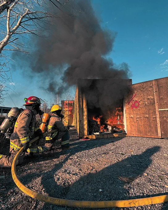 Two firefighters, wearing protective gear and helmets, are using a hose to extinguish a fire inside a burn cell. Thick black smoke is billowing from the opening. The scene is set outdoors on a gravel surface, with a clear blue sky in the background.
