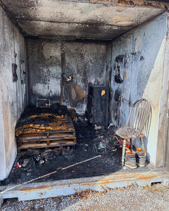 A small, charred room is shown inside a burn cell, visibly damaged by fire. Inside, there is a burnt bed made of wooden pallets, a scorched chair with a few items on it, and a partially intact shelf. The room's walls are blackened, and debris is scattered on the floor.

