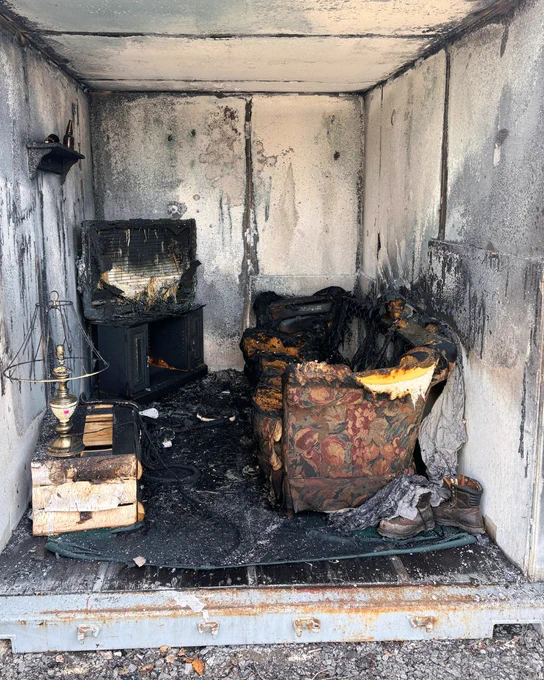 Inside a burn cell with severe fire damage. The interior is charred with soot-covered walls. Burned furniture, including a scorched television and a heavily damaged armchair, are visible. A singed lamp and some charred books are on a low table. Debris covers the floor.

