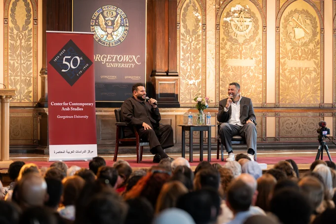 A discussion event at Georgetown University features two individuals seated on stage in front of an audience. A banner for the Center for Contemporary Arab Studies is displayed on the left. The background includes ornate wood paneling and the Georgetown University insignia.