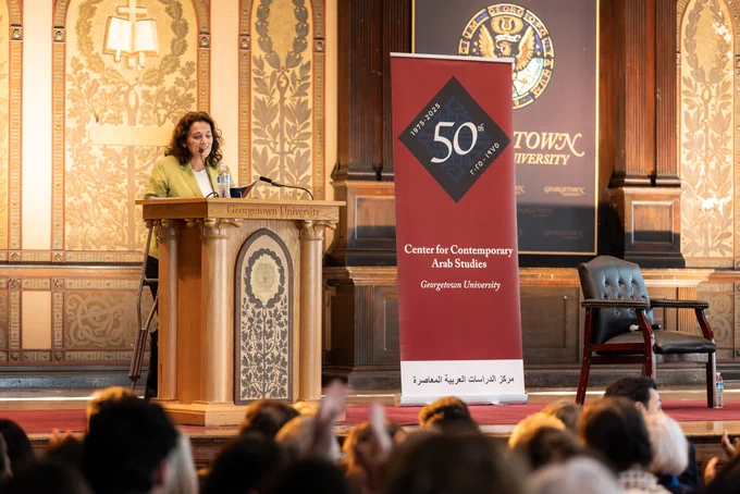 A person is speaking at a podium with a Georgetown University emblem in the background. The setting appears to be an auditorium. A banner next to the podium mentions the Center for Contemporary Arab Studies 50th anniversary. There is a seated audience visible in the foreground.