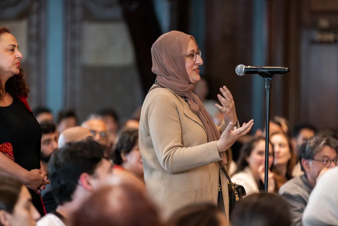 A person speaks into a microphone in a crowded room, gesturing with hands. The audience listens attentively.