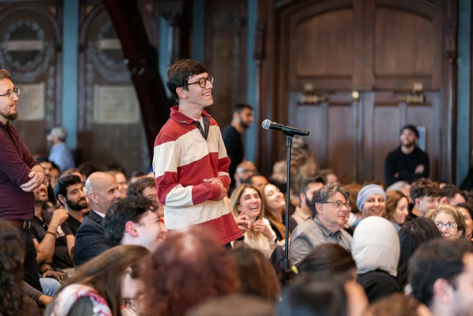 A person stands at a microphone, smiling, in a crowded room filled with seated attendees. The atmosphere seems lively and engaging, with people watching attentively.