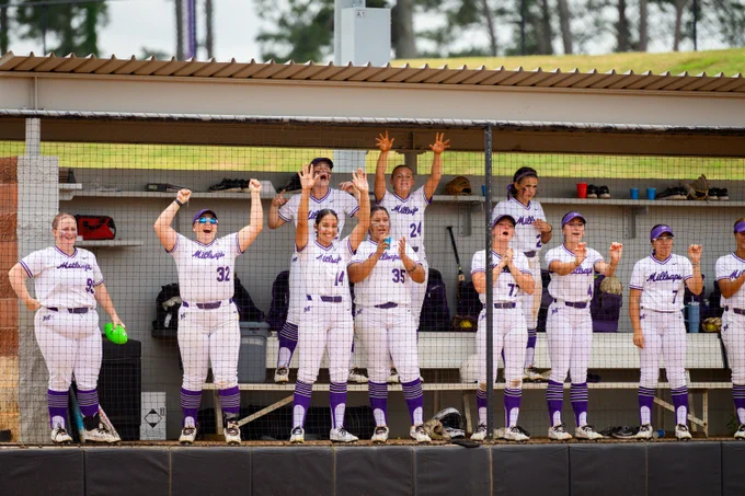 The image shows a group of Millsaps College softball players in purple and white uniforms, energetically cheering in a dugout during a game. Some players have their arms raised, others are clapping, and a few are sitting. The dugout is covered, with equipment and benches visible inside. The atmosphere appears lively and supportive.
