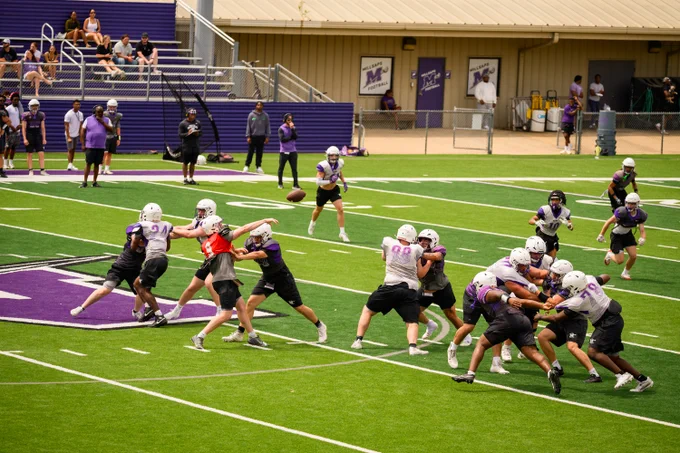 Millsaps College football team participating in our Purple & White Spring game on a field marked with a large "M". They are wearing protective gear and helmets. Several individuals observe from the bleachers in the background.