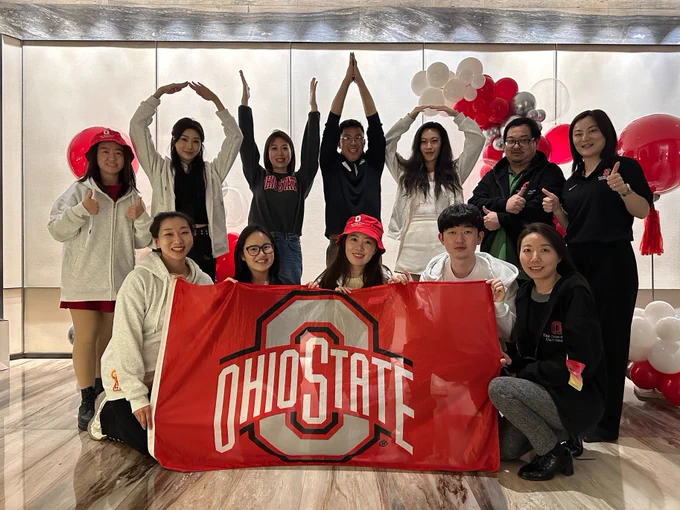 A group poses indoors with an "Ohio State" banner. Some are wearing Ohio State apparel. Red and white balloons decorate the background.