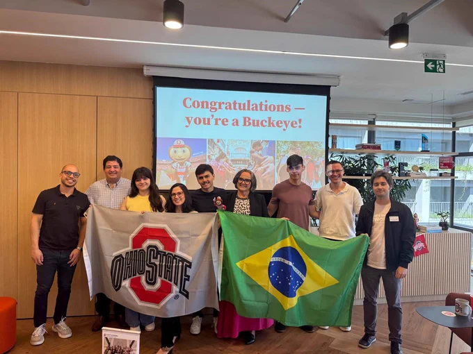 A group of people smiling indoors, with two holding flags: one of Ohio State and the other of Brazil. A screen behind them displays the message "Congratulations — you're a Buckeye!" with colorful images.