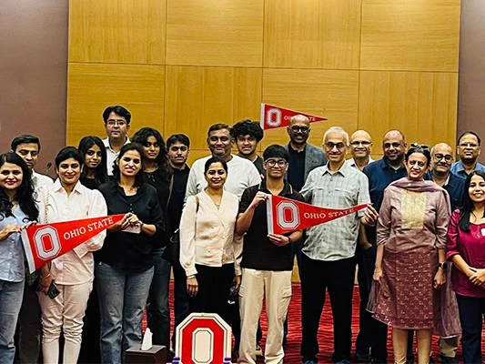 A group of people standing together in a room with wooden paneling and red carpet. Some individuals are holding Ohio State University banners and a logo cutout.