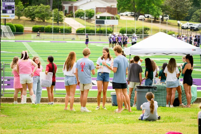 A group of people are gathered on a grassy area in front of the Millsaps College football field during our spring Purple & White day. Some are chatting near a white canopy, while others are facing the field. Players are seen in the background on the field, and there are trees and buildings in the distance.