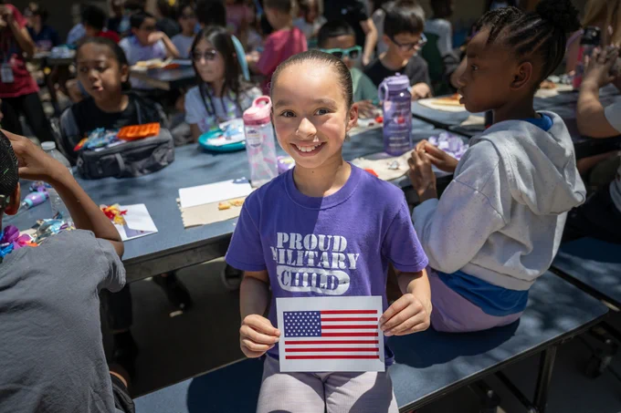 Child wearing a purple shirt that says 'Proud Military Child' holding an American flag card while sitting at a picnic table with other children outdoors.