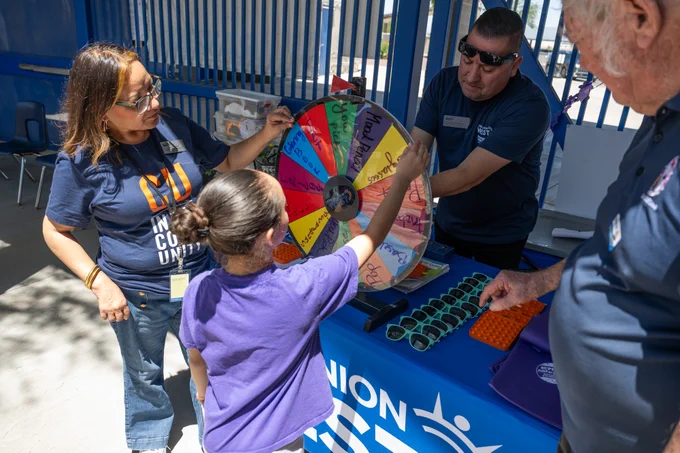 A young girl spins a colorful prize wheel at an outdoor event booth with three adults watching nearby.