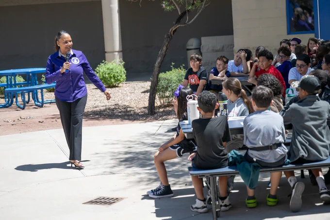 A woman in a purple jacket speaks into a microphone addressing a group of children seated outdoors at picnic tables on a sunny day.